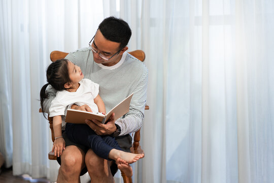 Asian father reading storybook to daughter in living room, family concept and early childhood development - Powered by Adobe