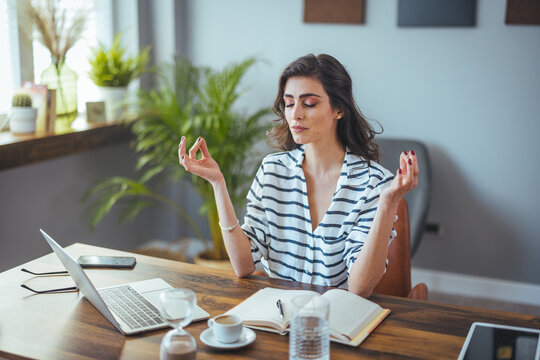 Young woman near the laptop, practicing meditation at the office desk, in front of laptop, online yoga classes, taking a break time for a minute, healing from paperwork and laptop radiation