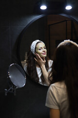 Fototapeta na wymiar Woman in headband applies green clay mask to her face in front of a mirror