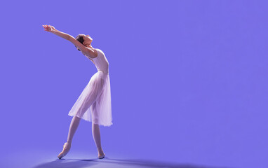 elegant ballerina in pointe shoes is dancing in a long flying white skirt on a lilac light...