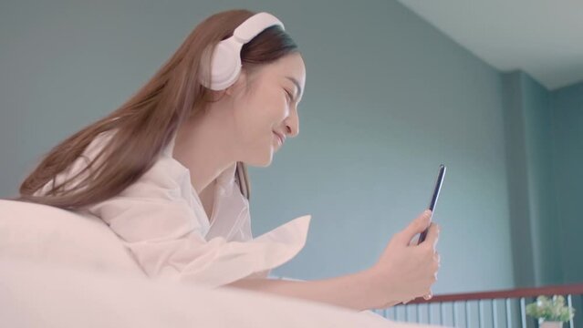 Beautiful Asian woman use mobile phone , video call and use headphone listen the  music while lying down on the bed. Young beautiful Asian woman use mobile phone. Lesbian couple relationship concept.