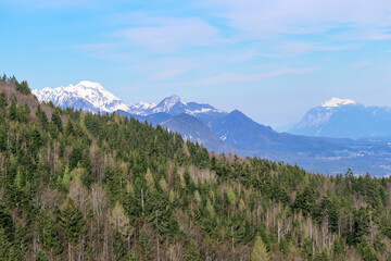 Scenic view of snow capped mountain peaks of Karawanks near Sinacher Gupf in Carinthia, Austria. Mount Mittagskogel and Dobratsch is visible through dense forest in early spring. Sunny Rosental