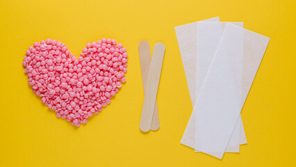 Pink heart-shaped wax granules for depilation, strips for depilation and wooden spatulas on a yellow background. Epilation, depilation, removal of unwanted hair with wax. Top view. - obrazy, fototapety, plakaty