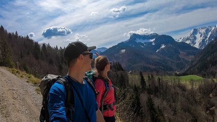 Fototapeta na wymiar Couple with backpack and scenic view of snow capped mountain peaks of Karawanks near Sinacher Gupf in Carinthia, Austria. Mount Hochstuhl (Stol) visible through forest in spring. Rosental sunny day
