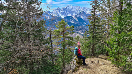 Naklejka na ściany i meble Woman with backpack sitting on a rock with scenic view of snow capped mountain peaks of Karawanks near Sinacher Gupf in Carinthia, Austria. Mount Wertatscha is visible through dense forest in spring