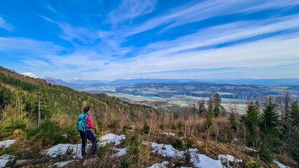 Woman with backpack and a scenic view on the Drava river in the Rosental valley on the way to Sinacher Gupf in Carinthia, Austria. Forest in early spring. Austrian Alps mountain ranges on sunny day