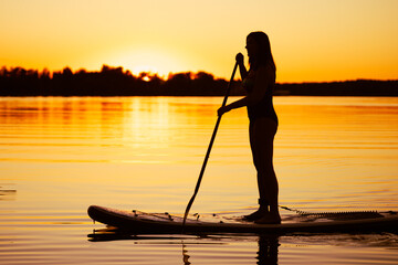 Silhouette of middle aged Caucasian woman standing on sup board with oar in hands on lake in evening with magnificent sunset in background. Active lifestyle. - obrazy, fototapety, plakaty