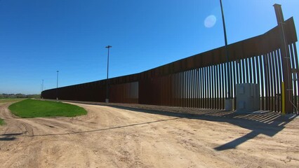 Rear POV driving on service road away from the border wall between USA and Mexico; near McCallen Texas on a sunny day; concepts of national defense, border security, and Trump's build the wall