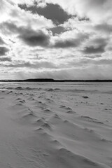 Black and white photo of patterns in the snow on the shore of Grand Traverse Bay with Power Island (aka Marion Island) in the distance under a cloudy sky.