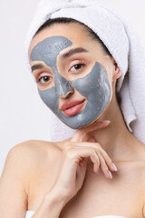 A woman with a gray clay cosmetic mask in a white towel on her head on a white background.