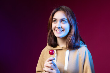 Portrait of a woman in a hoodie, girls licking a red round lollipop with beautiful makeup on a purple trendy background in the studio.