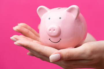 A cute girl in a pink jacket is holding a 3d piggy bank in her hands. Beautiful close-up portrait in studio on pink.