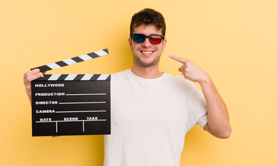 young handsome man smiling confidently pointing to own broad smile. cinema clapper concept