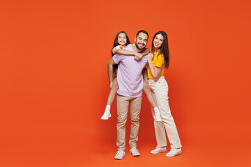 Fototapeta premium Full size young fun happy parents mom dad with child kid daughter teen girl in basic t-shirts giving piggyback to daughter isolated on yellow background studio Family day parenthood childhood concept