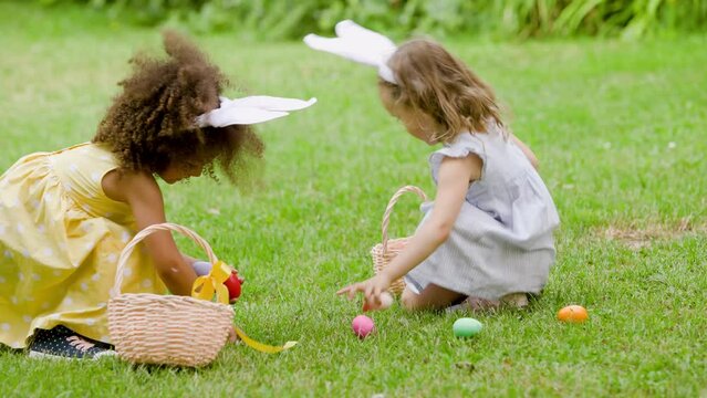 two girls are sitting on the green lawn during easter egg hunt . Child wear pink rabbit ear and putting Easter eggs in to baskets
