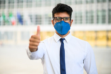 Young businessman outdoor in a modern setting giving thumbs up and wearing a protective mask...