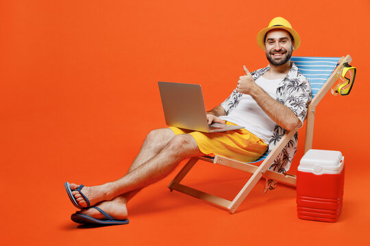 Young Tourist Man Wear Beach Shirt Hat Lie On Deckchair Hold Use Work On Laptop Pc Computer Show Thumb Up Isolated On Plain Orange Background Studio Portrait. Summer Vacation Sea Rest Sun Tan Concept