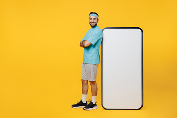 Full body young fitness trainer instructor sporty man sportsman in headband blue t-shirt big screen mobile cell phone with workspace mockup isolated on plain yellow background. Workout sport concept.