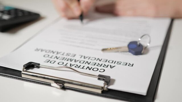Standard Residential Lease Agreement in Spanish speaking language. Female or male hands sign documents. In the foreground lies the key to the apartment or house.