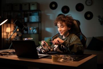 Boy sits at desk in room in evening, finishes soldering robot which is school project, child watches tutorials, instructions on laptop, learns step by step to fix electronics