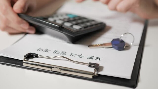 Standard Residential Lease Agreement in Korean speaking language. The hands of realtor calculate the cost on calculator. In the foreground lies the key to the apartment or house.