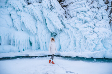 Slender girl traveler in winter clothes on the background of ice rocks. Nature of Lake Baikal in winter. Winter landscapes. White winter jacket, red boots. Blue ice.