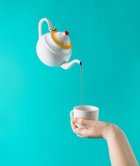 A white teapot pours tea into a cup that is in his hand on a turquoise blue background. Minimal layout composition.