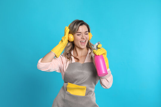 Beautiful young woman with headphones and bottle of detergent singing on light blue background
