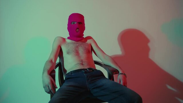 Anonymous shirtless man in balaclava sitting in chair near wall. Unrecognizable dangerous male with naked torso in pink balaclava posing.