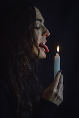 Playing with fire - young beautiful woman with long hair in profile holding a burning candle in her hand and pretending to lick its flame