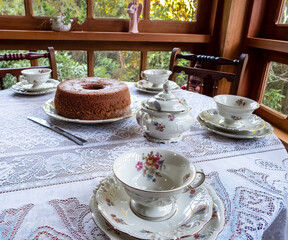 Brown cake on a wooden table with white KPM porcelain tea set, decorared with flowers. White lace tablecloth, silver knife,  tea cup, saucer, plate, sugar pot. 