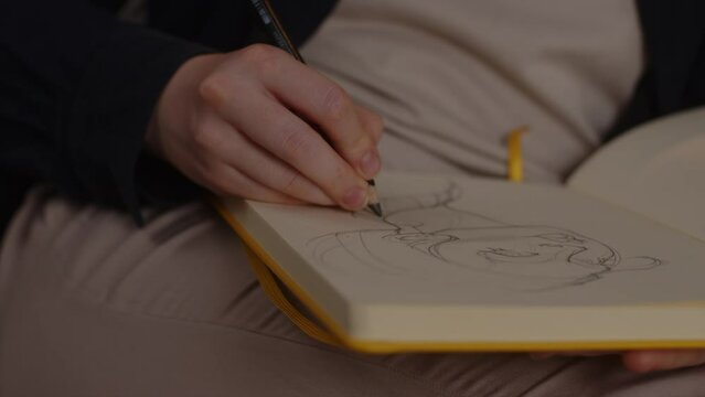 The artist draws a sketch of a girl in a yellow paper sketchbook sitting on a sofa in the living room