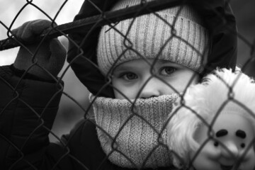 Fototapeta na wymiar A little refugee girl with a sad look behind a metal fence. Social problem of refugees and internally displaced persons. Russia's war against the Ukrainian people