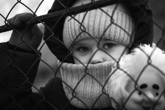 A little refugee girl with a sad look behind a metal fence. Social problem of refugees and internally displaced persons. Russia's war against the Ukrainian people