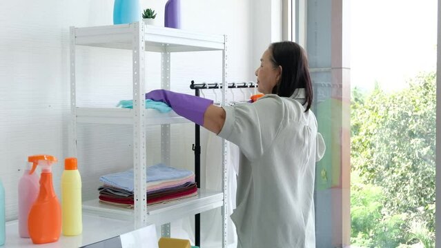 Senior Asian woman, housewife cleaning books on bookshelf with feather duster in living room.
