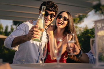 Couple of sweethearts standing on the table pouring sparkling wine champagne into their friends...