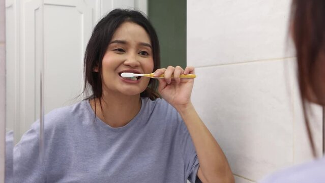 Smiling young woman with toothbrush cleaning teeth and looking mirror in the bathroom.