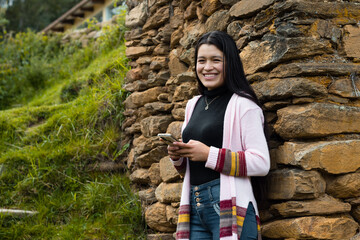 Una atractiva joven sonriente con el pelo largo y negro parada cerca de una pared de roca con su móvil en las manos - obrazy, fototapety, plakaty