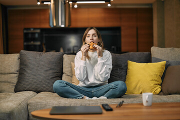 Young woman eating burger junk food watching tv series or movie, sitting on sofa in living room,...