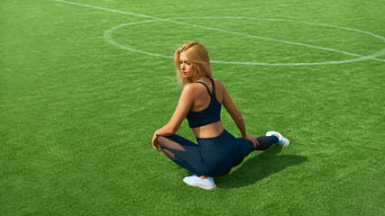 Outdoor fitness. Attractive sportswoman preparing for sports. Fitness female sports model doing stretching on a green grass football field on a sunny day. The athlete turned her back to the camera