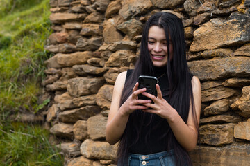 Una joven sonriente con el pelo largo y negro parada cerca de una pared de roca mirando  su móvil en las manos - obrazy, fototapety, plakaty