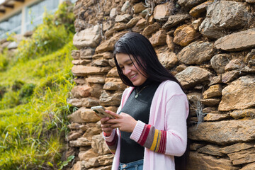 Una atractiva joven sonriente con el pelo largo y negro parada cerca de una pared de roca mirando  su móvil en las manos - obrazy, fototapety, plakaty