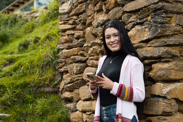 Una joven sonriente con el pelo largo y negro parada cerca de una pared de roca con su móvil en las manos - obrazy, fototapety, plakaty