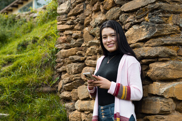 Una atractiva joven con el pelo largo y negro parada cerca de una pared de roca con su móvil en las manos - obrazy, fototapety, plakaty