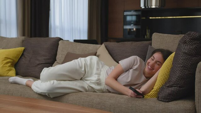 Laughing happy beautiful young girl watching sitcom movie or stand up comedy, lying on sofa, relaxing at home