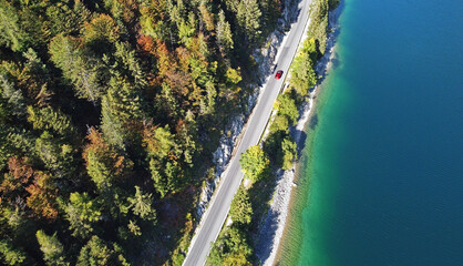 Luftaufnahme mit einer Drohne von einer Straße mit rotem Auto zwischen einem See und dem...