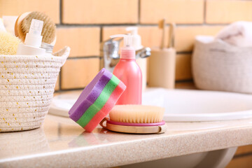 Massage brush, bath sponge and bottle of cosmetic product on table near sink