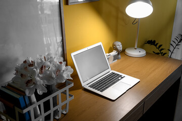 Modern laptop and glowing lamp on desk near yellow wall