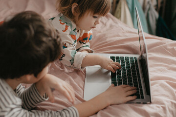 Little brother and sister lie in the bedroom on the bed and look at the laptop