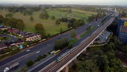 Subway metro commuter train arriving elevated station platform on track, passenger train aerial drone cityscape with four lane road traffic, apartment construction and parkland in distance